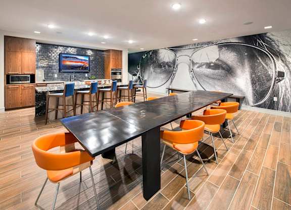 A black and white photo of a woman's face is on the wall behind a long black table with orange chairs.