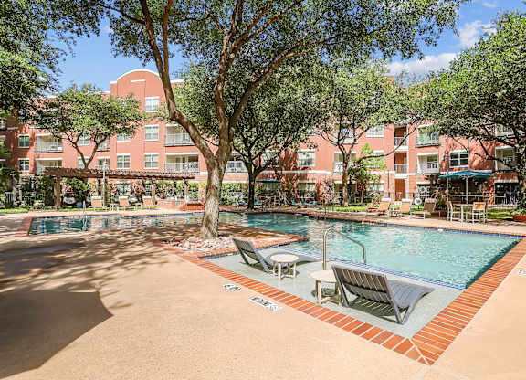 A pool surrounded by trees and chairs in front of a red building.