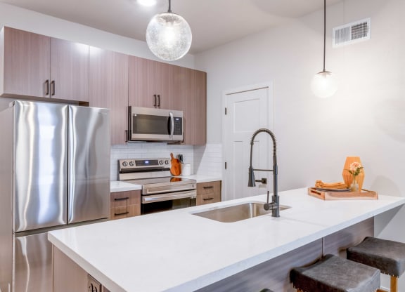 2-Bedroom Apartments in Raymore, MO - The Venue Townhomes - Designer Kitchen with Island, Pantry Stainless Steel Samsung Appliances, Wood-Style Cabinetry, White Tile Backsplash, White Quartz Countertops, Bar-Stool Seating, and Pendant Lights.