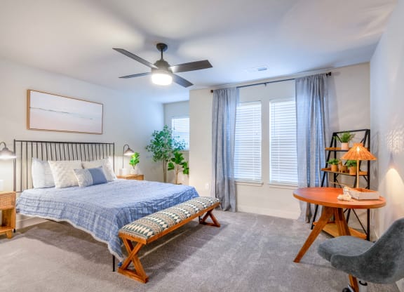 Two-Bedroom Apartments in Raymore, MO - The Venue Townhomes - Bedroom with Carpeted Flooring, Bed, Footboard Bench, Wooden Nightstands with Lamps, House Plants, Circular Wooden Table, Grey Office Chair, Ceiling Fan, Display Shelf, and Large Windows.