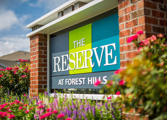 A sign for The Reserve at Forest Hills is shown in front of a brick wall and flowers.