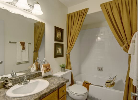 Elegant bathroom here at Desert Horizon Apartments with beige tones. Features a granite countertop, white sink, and tub with golden curtains. Decor includes framed art and folded towels.