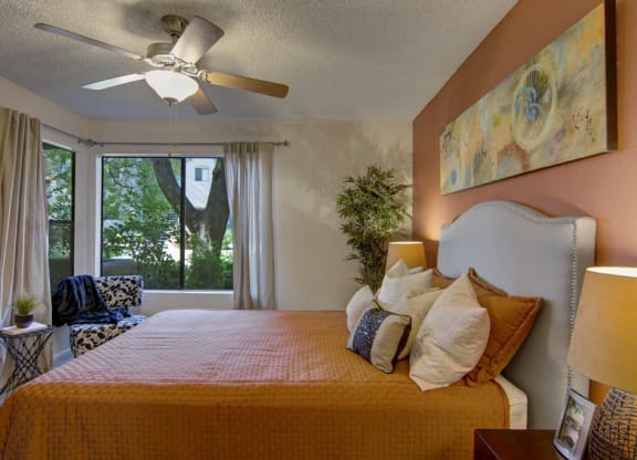 Cozy bedroom here at Desert Horizon Apartments with brown and cream decor, a large bed with many pillows, abstract art on a warm accent wall, and a window with tree view.