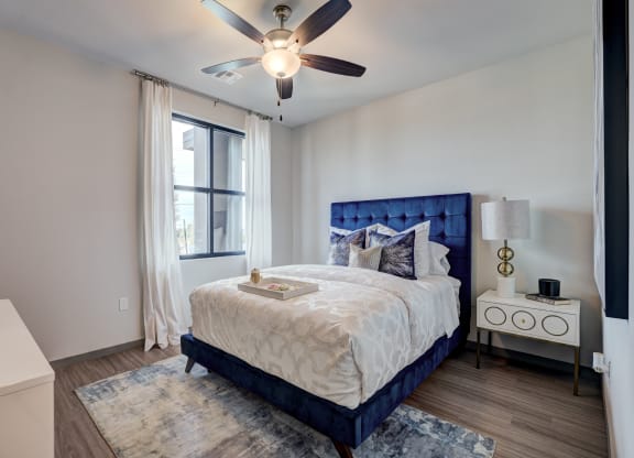 Cozy bedroom here at 35 Degrees North Apartment Homes with a blue tufted bed, white bedding, and decorative pillows. A tray sits on the bed. A lamp and decor adorn the nightstand. Soft, natural light filters through the window, creating a serene ambiance.