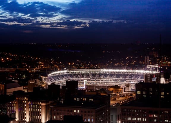 A brightly lit baseball stadium near 4Marq Apartments at night, bustling with fans. Surrounding city lights twinkle under a deep blue sky with scattered clouds. Electric atmosphere.