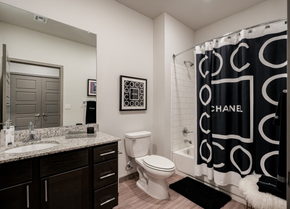 Modern bathroom here at 35 Degrees North Apartment Homes with a granite-topped vanity, large mirror, and white toilet. A black-and-white Chanel-themed shower curtain adds elegance.