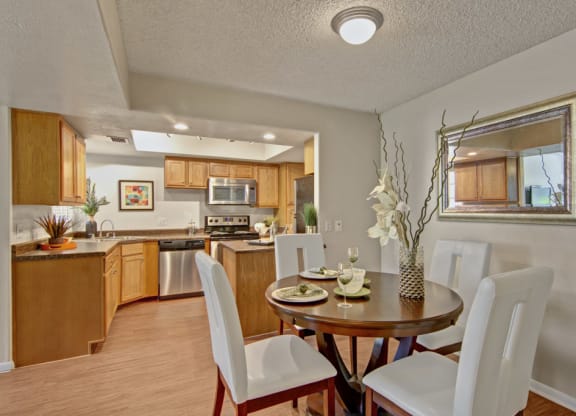 Dining area here at Desert Horizon Apartments with a round wooden table set for four, white chairs, and a vase with flowers. Adjacent modern kitchen with wood cabinets and stainless steel appliances.