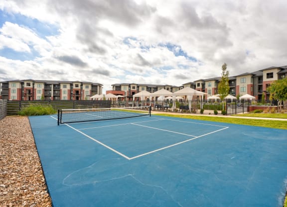 A blue tennis court here at 35 Degrees North Apartment Homes under a partly cloudy sky with modern apartment buildings in the background. Nearby, white canopies cover outdoor seating areas.