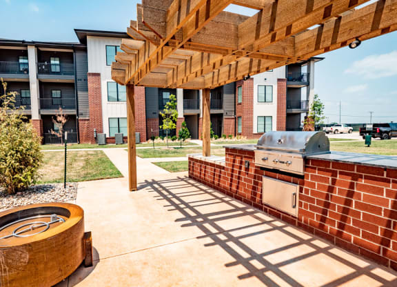 Outdoor BBQ area here at 35 Degrees North Apartment Homes with a brick grill under a wooden pergola. Nearby is a modern fire pit. The background shows a multi-story apartment building.
