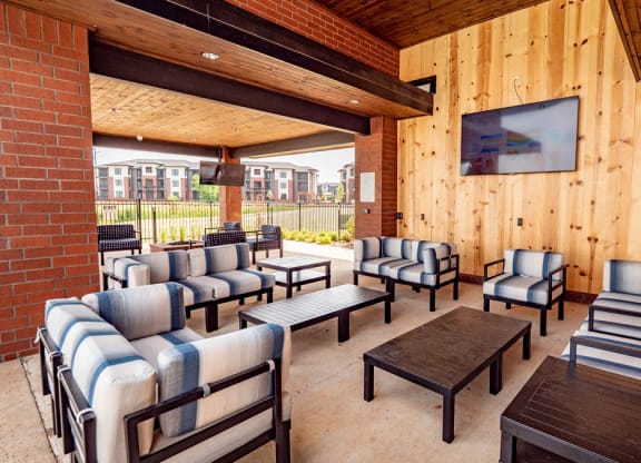 Covered patio here at 35 Degrees North Apartment Homes with striped cushioned seating, wooden ceiling, and brick walls. A large TV is mounted on the wooden wall. Apartments are visible outside.