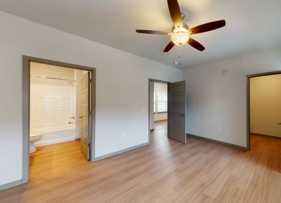 Empty room here at 35 Degrees North Apartment Homes with wood floors, gray doors, and a ceiling fan. Open doors reveal a bright bathroom with white tiles and a window-lit adjacent room.