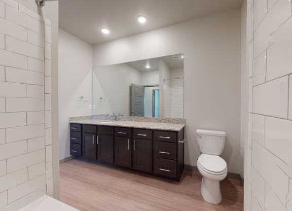 Modern bathroom here at 35 Degrees North Apartment Homes with white subway tile, large mirror, granite countertop, dark wood cabinets, and a white toilet. Calm and clean ambiance.