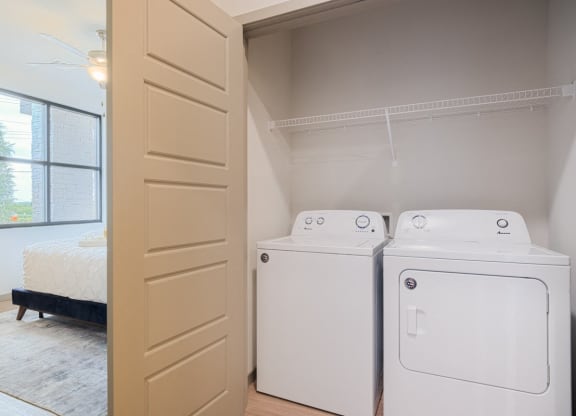 Laundry area here at 35 Degrees North Apartment Homes with a white washer and dryer in a closet. Beige sliding door partially open, revealing a bright bedroom with a bed and large window. Calm and tidy atmosphere.