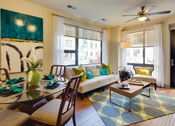 Modern living room here at Inspire Apartment Homes with a beige sofa adorned with green and turquoise pillows, abstract art, a glass dining table, and a geometric rug. Bright and inviting.