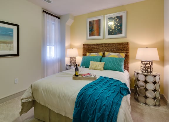 Cozy bedroom here at Inspire Apartment Homes with a wicker headboard, teal bedding, and yellow accents. Soft lighting from modern lamps, abstract art, and a tray with wine add elegance.