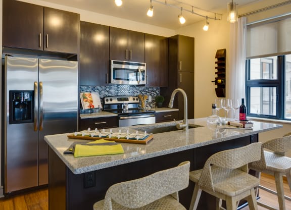 Modern kitchen here at Inspire Apartment Homes with dark wood cabinets, stainless steel appliances, and a granite island with wicker stools. Warm lighting and a cozy ambiance.