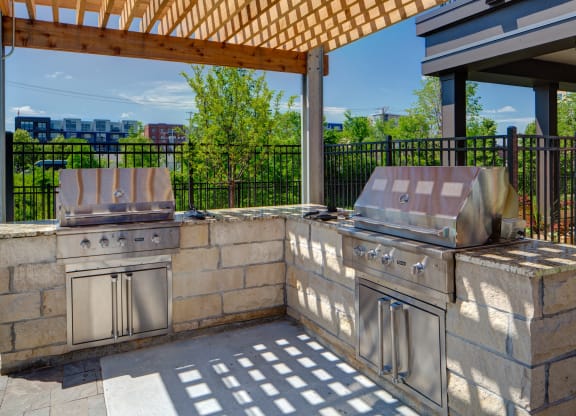 Outdoor patio here at Inspire Apartment Homes with two stainless steel grills on a stone counter under a wooden pergola. Shadow patterns on the ground and sunny greenery in the background.