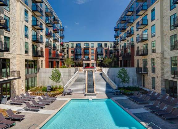 Modern Inspire Apartment Homes complex with a central pool and surrounding sun loungers. Flanked by two five-story buildings, a staircase leads to a rooftop terrace.