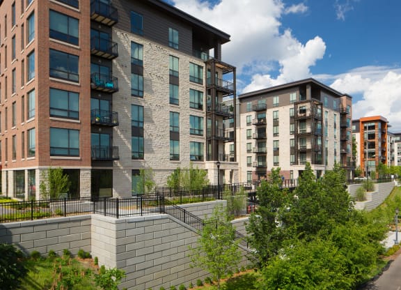 Modern Inspire Apartment Homes buildings with brick and glass facades are surrounded by lush greenery under a bright blue sky with scattered clouds.