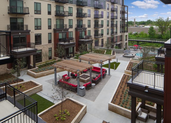 Modern courtyard here at Inspire Apartment Homes with a symmetrical design featuring wooden pergolas, red sofas around fire pits, lush landscaping, and a distant pool area.