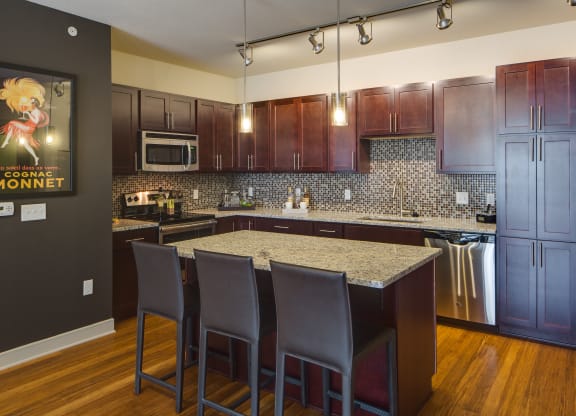 Modern kitchen here at Inspire Apartment Homes with dark wood cabinets, granite countertops, and mosaic backsplash. An island with three brown chairs and stainless steel appliances. Hanging lights create a cozy ambiance.