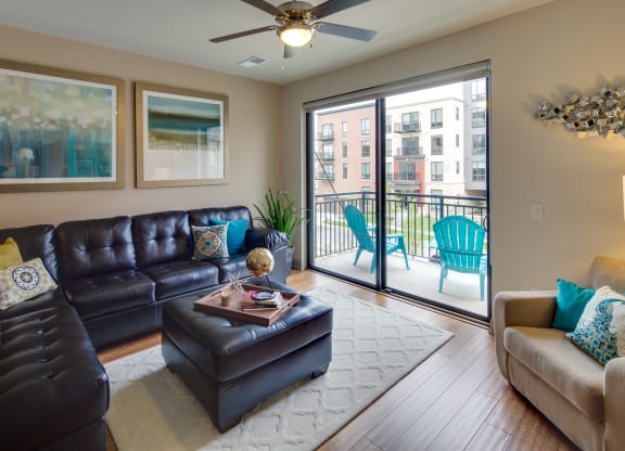 Modern living room here at Inspire Apartment Homes with a black leather sectional, colorful cushions, large abstract art, and sliding doors leading to a balcony with blue chairs. Cozy ambiance.