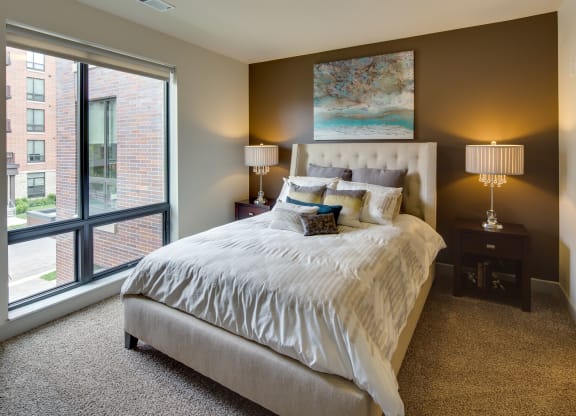 Elegant bedroom here at Inspire Apartment Homes with a plush bed and assorted pillows, flanked by bedside tables and lamps. Large window shows a cityscape, creating a cozy ambiance.