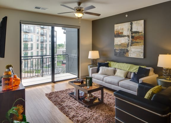 Modern living room here at Inspire Apartment Homes with a gray accent wall, abstract painting, and a white sofa adorned with colorful cushions. Sliding glass doors lead to a balcony with a cityscape view. A ceiling fan and warm lighting create a cozy ambiance.
