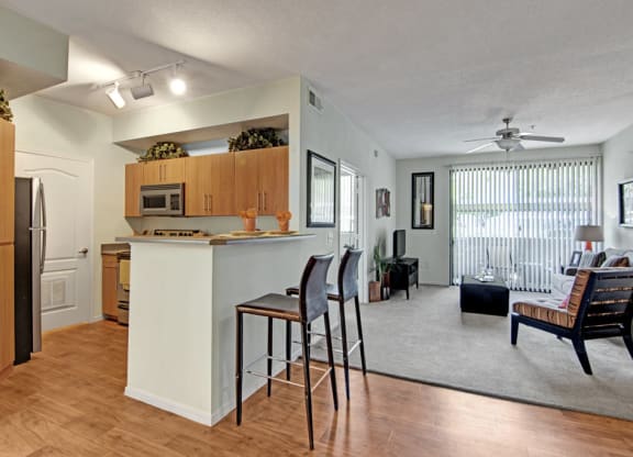 Modern open-plan living room and kitchen here at Acclaim Apartments with wooden flooring, light walls, and neutral decor. A countertop with bar stools divides the spaces. A ceiling fan and large window with vertical blinds add to the bright, airy feel.