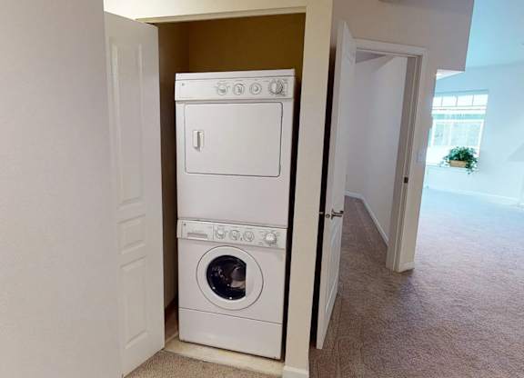 A hallway laundry closet at Admirals Cove featuring a stacked washer and dryer set behind a partially open door. Soft neutral carpet and nearby bedroom and living room entrances create an easy flow through the home, offering convenient in-unit laundry access.