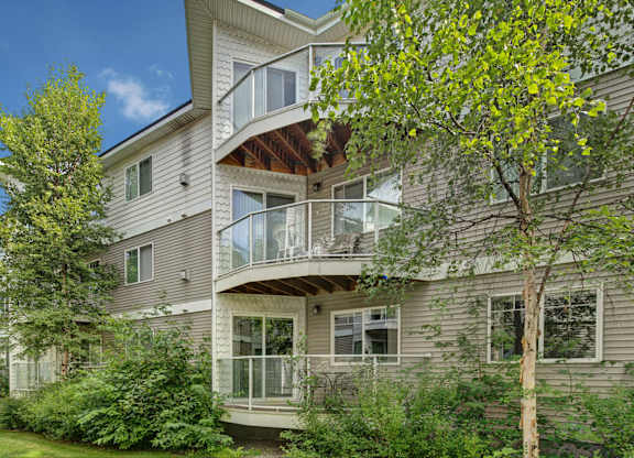 A three-story exterior view of Admirals Cove showcasing private balconies with glass railings surrounded by tall trees and green landscaping. Light siding and shingle detailing give the building a clean, modern look. Outdoor seating on some balconies highlights a relaxing residential setting.