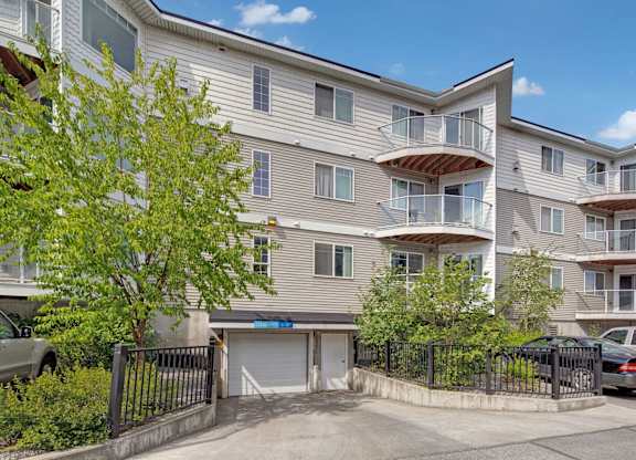 A multi-story exterior view of Admirals Cove featuring light siding, private balconies with glass railings, and landscaped greenery along the front. Parking is visible near the entrance, and a gated driveway leads to underground garage access beneath the building, showcasing convenient resident amenities.