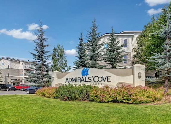 The entrance sign for Admirals Cove Apartment Homes set against manicured landscaping and tall evergreen trees. Buildings with balconies rise in the background, while a clear blue sky completes the welcoming community scene.