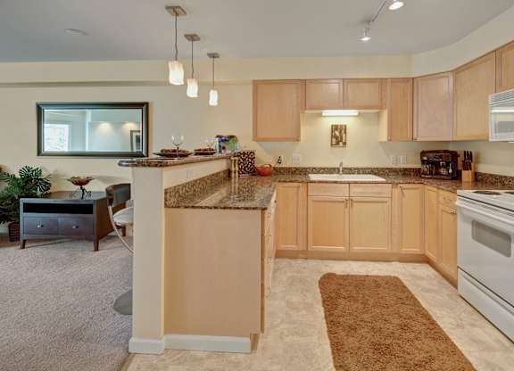 An alternate angle of a spacious kitchen here at Admirals Cove with light brown cabinets, granite counter tops, bar type seating, and a ceramic sink. The room has a warm tone.