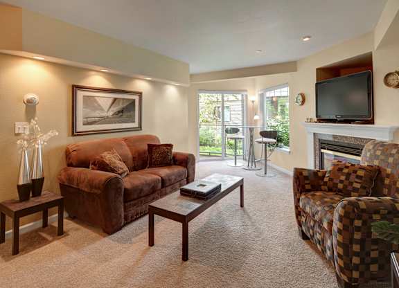 A cozy living room here at Admirals Cove with a light brown carpet and soft lighting. There is a brown couch, and a brown, checkered patterned sofa chair. There is a two-person table near a sliding glass door that leads you outside.