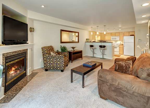 A warm and inviting living room at Admirals Cove featuring plush beige carpet and a cozy brown sofa beside a patterned armchair. A gas fireplace with a mounted TV adds comfort, while a large wall mirror opens the space visually. The room flows into a bright kitchen with light wood cabinets, white appliances, and a breakfast bar with two modern barstools.