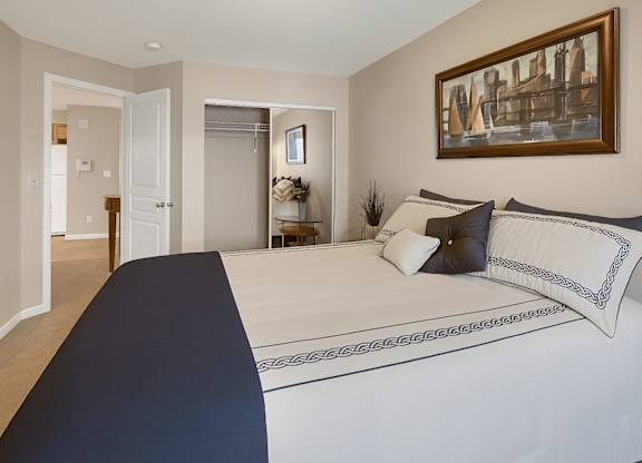 A bright bedroom at Admirals Cove featuring a neatly made bed with decorative pillows and patterned bedding. Soft beige walls and carpet create a warm feel. A sliding-door closet offers storage, and a doorway opens to the living area, giving the space an open, welcoming flow.