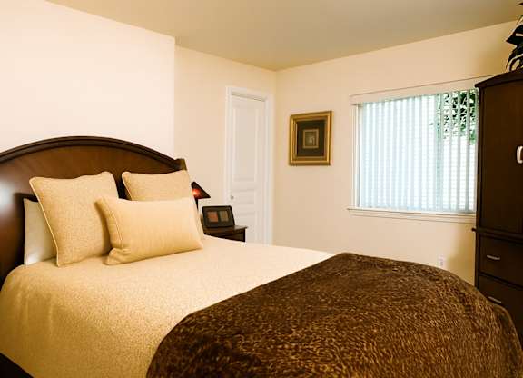 A cozy bedroom at Admirals Cove with warm cream walls and soft natural light filtering through window blinds. The room features a neatly made bed with plush pillows and a dark wood headboard, complemented by matching furniture pieces and simple wall art for a comfortable, inviting feel.