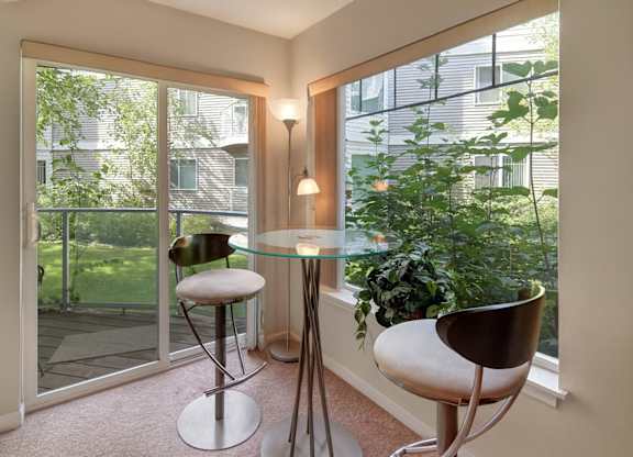 A bright dining nook at Admirals Cove featuring a round glass table with two modern bar-height chairs. Large windows and a sliding glass door overlook green landscaping, filling the space with natural light. Soft carpeting and corner lighting create a relaxed spot for meals or morning coffee.