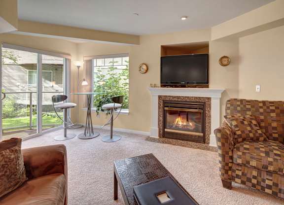 A bright living room at Admirals Cove with soft carpet flooring and a cozy seating area. A fireplace with a TV above serves as the focal point, while large windows and a sliding door bring in natural light. A small glass-top dining set sits beside greenery just outside, creating a relaxing indoor-outdoor feel.