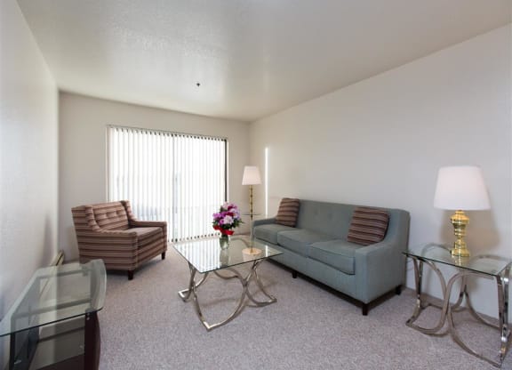 A modern living room here at Outlook Apartments with a gray sofa, striped armchair, glass coffee table, and two glass side tables with lamps. A vase with flowers adds color.