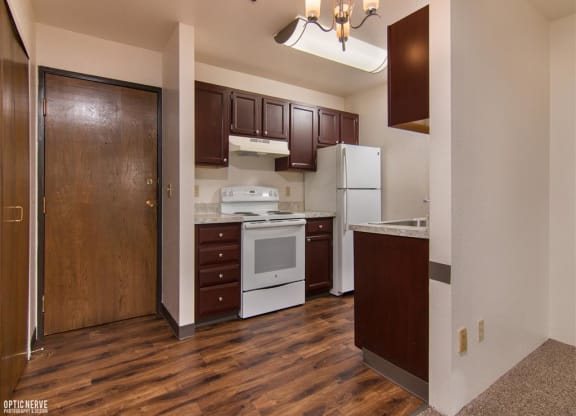 Compact kitchen here at Outlook Apartments with dark wood cabinets, white appliances, and marble countertops. A wooden door and elegant light fixture add warmth and character.