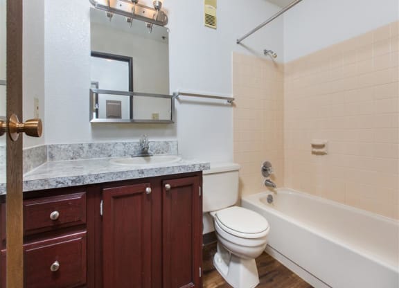 Compact bathroom here at Outlook Apartments with granite countertop, wooden cabinets, and a wall-mounted mirror. Beige tile surrounds the bathtub and shower, creating a clean, simple look.