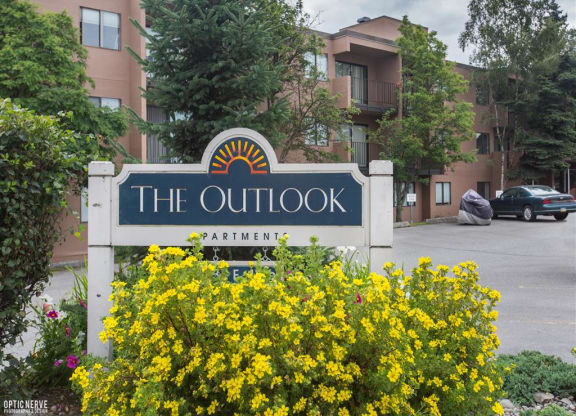 Sign reading "The Outlook Apartments" with a sun icon. Surrounded by vibrant yellow flowers, trees, and a brown apartment building in the background. Cozy atmosphere.