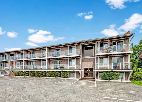 Exterior view of Arbor Pointe apartment building with balconies, large windows, and a parking lot in front under a blue sky with scattered clouds.