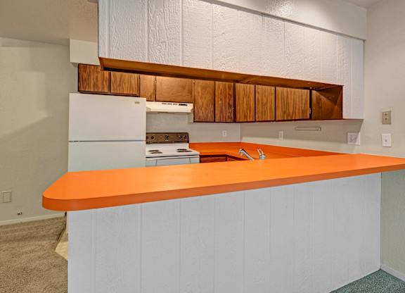 Close-up view of an Arbor Pointe kitchen with a long bright orange countertop, white base cabinets, wood upper cabinets, and white appliances including a stove and refrigerator.