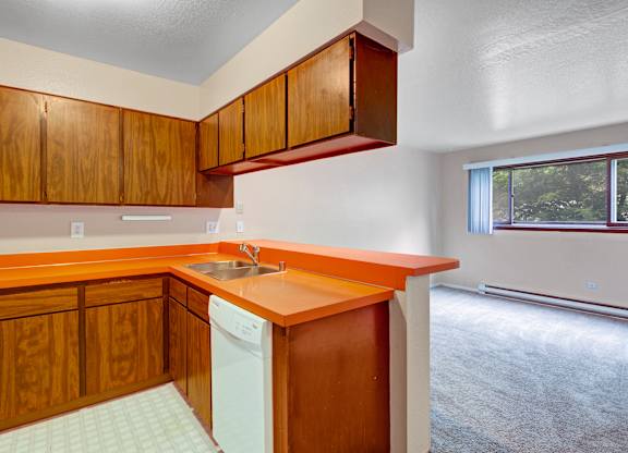 Kitchen with orange countertops and wood cabinets next to a carpeted living room with a large window letting in natural light.