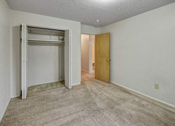 A bright bedroom at Arctic Sun Apartments with plush beige carpet and neutral walls. An open closet with double doors offers storage space. A doorway leads to the hallway and bathroom, and a ceiling light adds soft illumination, creating a comfortable and versatile room layout.