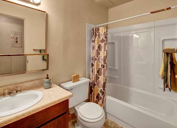 A warm bathroom at Arctic Sun Apartments with a tub and shower combo and a patterned curtain. The space includes a vanity with a sink, a wide mirror, and storage below, along with a toilet. Soft lighting and neutral tones create a comfortable, practical layout.