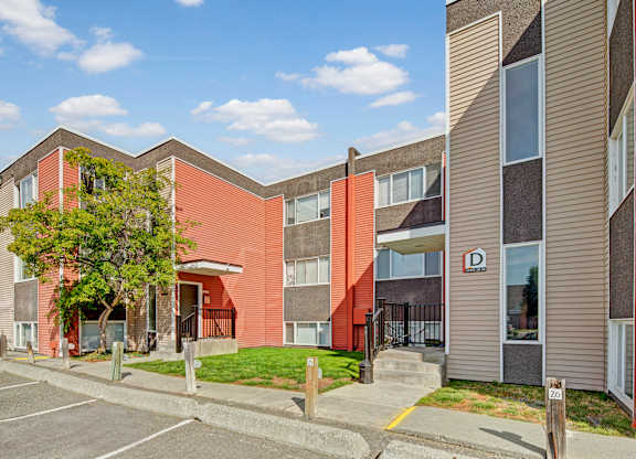 A sunny exterior view of Arctic Sun Apartments showing a two-story building with red and tan siding, large windows, and secure entryways. Small lawns and trees add greenery, and numbered parking spaces sit right outside for resident convenience. The sky is bright with scattered clouds.